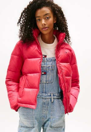 Rode pufferjack met een hoge kraag en ritssluiting, met een glanzende textuur. Draagt over blauwe denim overalls met voorzakken.