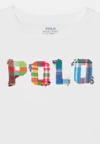 Vit T-shirt med "POLO" i flerfärgad rutigt tygbuchstäver, prydd med små pärlor längs kanterna på bokstäverna.
