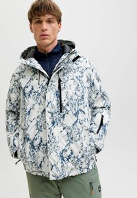 Veste à motifs blanche et marine avec capuche, dotée de zips, d'un col montant et de multiples poches, fabriquée en matériau résistant à l'eau.