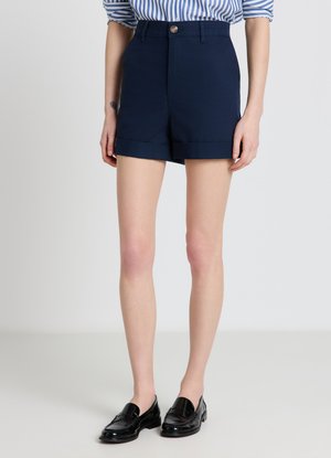 Femme portant un short taille haute bleu marine avec revers, une chemise rayée bleue et blanche aux manches retroussées, et des mocassins en cuir noir.
