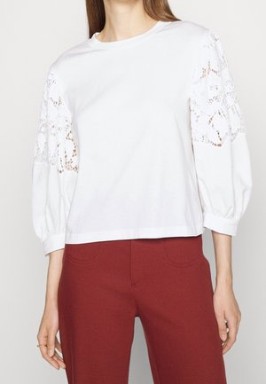 Donna che indossa una camicetta bianca con maniche floreali in pizzo e pantaloni a vita alta color ruggine, in piedi davanti a uno sfondo semplice.