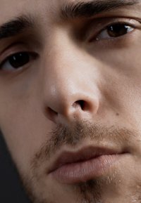 Gros plan sur un visage masculin avec une peau claire, une barbe de trois jours, des pommettes bien définies et des yeux brun foncé avec des reflets subtils et une texture.