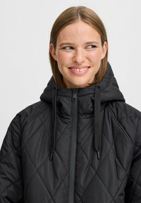 Veste noire matelassée avec un col haut, fermeture éclair et capuche réglable avec cordon. Présente des motifs en losange et un tissu texturé.