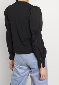 Donna in una blusa nera a maniche lunghe e con maniche a sbuffo, dotata di polsini abbottonati, e pantaloni in velluto a coste azzurri chiari, in posa su uno sfondo neutro.
