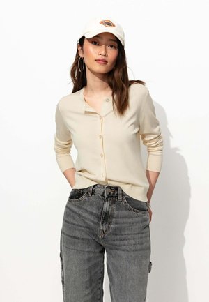 Gilet crème à boutons, à manches longues, associé à un jean en denim foncé taille haute. Mannequin portant une casquette blanche avec un logo orange.