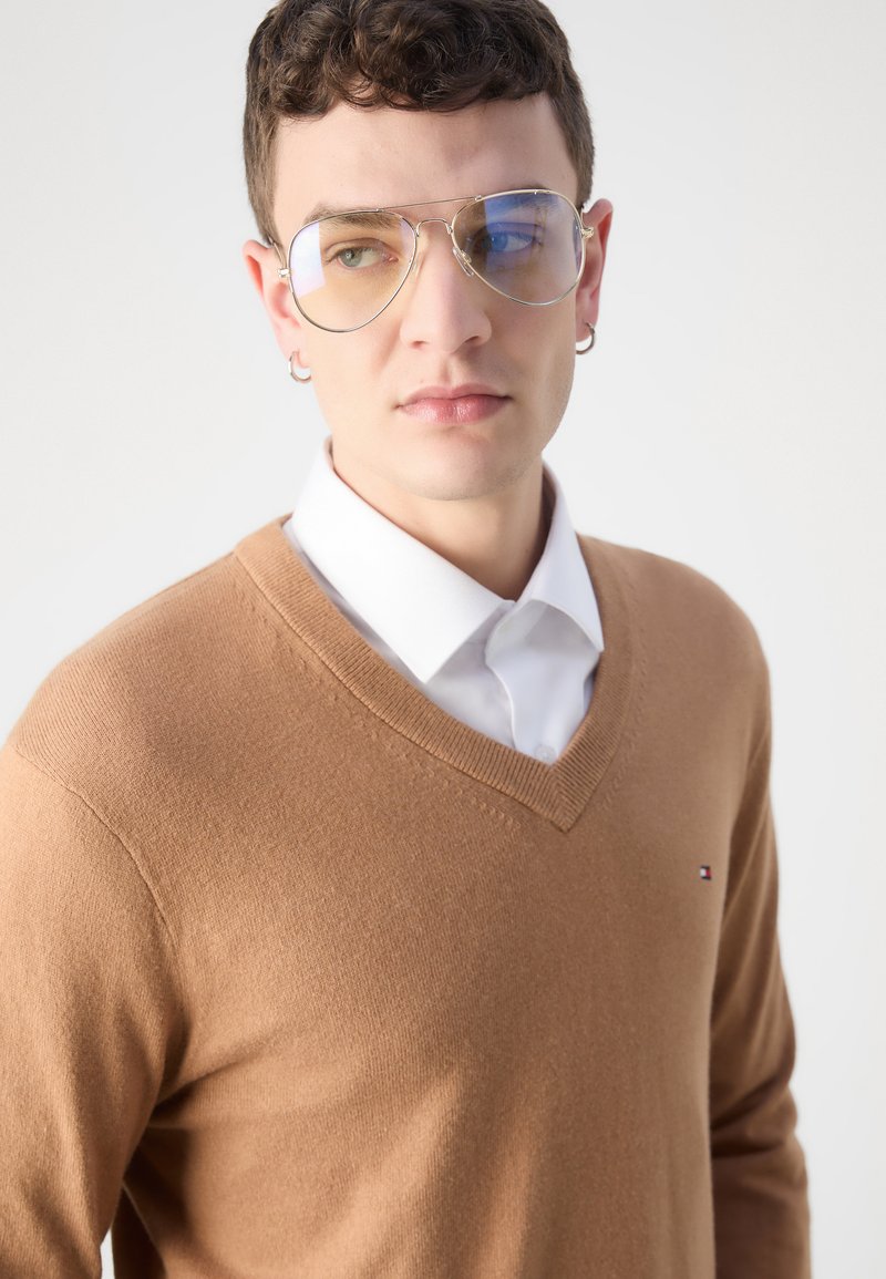 Maglione V a collo marrone, tessuto lavorato a maglia, indossato sopra una camicia bianca con colletto. Occhiali da sole grandi e rotondi con lenti blu. Logo distintivo esposto.