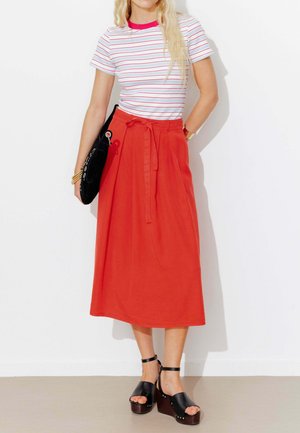 Femme portant un t-shirt blanc et rouge à rayures, une jupe midi rouge taille haute avec ceinture, des sandales compensées noires, et tenant une pochette noire.