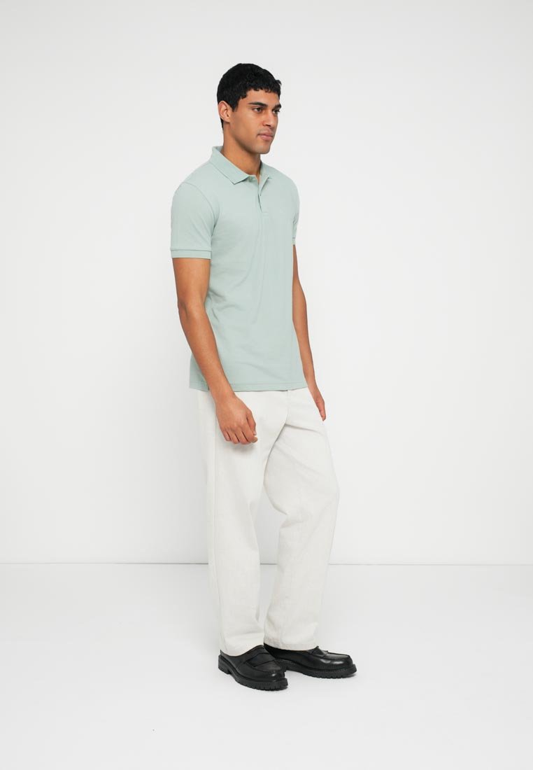 Polo vert clair à col, manches courtes, associé à un pantalon blanc à jambes larges et des chaussures noires, sur un fond uni.