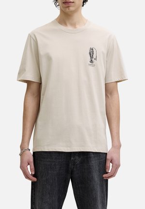 Jeune homme portant un t-shirt beige avec un petit graphisme noir de homard et le texte « Hooked on Adventure » sur la poitrine gauche, associé à un jean foncé.
