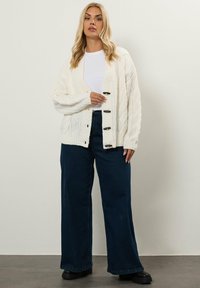 Kvinna med långt blont hår som bär en vit stickad kofta över en vit skjorta, mörka vida jeans och svarta stövlar, stående mot en vanlig vägg.