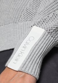 Szary sweter dzianinowy z fakturowanym wzorem na korpusie, ściągaczami oraz białą metką z logo "Calvin Klein".