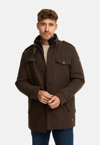 Brun uldfrakke med høj krave, knapper foran og lommer med klapper. Har skulderstropper og en glat tekstur. Kombineret med en lys sweater.