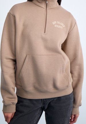 Beige sweatshirt med kvart-lang lynlås og kængurulomme, med teksten "New Balance Athletics", båret sammen med mørke jeans.