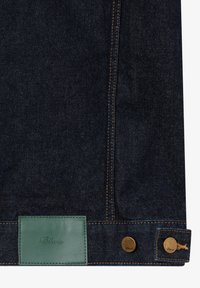 Dunkelblauer Denim-Stoff mit orangefarbenen Nähten, versehen mit einem grünen Lederpatch und zwei Messingknöpfen, die mit dem Markennamen geprägt sind.