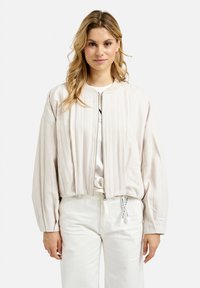 Lightweight, cropped bomber jacket in cream fabric with a ribbed texture, zip closure, and voluminous sleeves. White pants with drawstring.