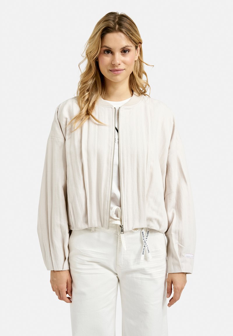 Lightweight, cropped bomber jacket in cream fabric with a ribbed texture, zip closure, and voluminous sleeves. White pants with drawstring.