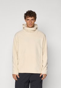 Beige fleece pullover with a high collar, front pocket, and long sleeves. Soft texture with stitching details. Dark pants not shown.