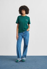 Groen katoenen T-shirt met een klein roze logo, gecombineerd met lichtblauwe wijde jeans en groene sokken. Effen achtergrond.