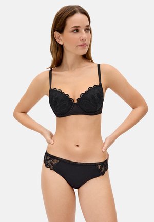 Mujer con sujetador negro de encaje y bragas a juego con detalles de encaje, de pie con las manos en las caderas, mirando de lado contra un fondo liso.