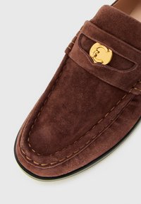 Brown suede loafer with visible stitching and gold circular metal buckle detail on the upper front area.