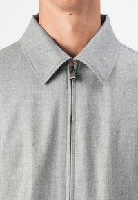 Gray zip-up jacket with a broad collar, textured fabric, and subtle detailing around the zipper. Smooth, straight design.