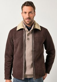 Hombre con barba que lleva una chaqueta de shearling marrón sobre un suéter de punto beige con botones y jeans azules, de pie contra un fondo liso.