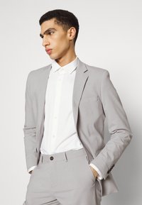 Completo grigio chiaro con una vestibilità slim, giacca monopetto, revers a lancia e pantaloni coordinati, abbinato a una camicia bianca con bottoni.