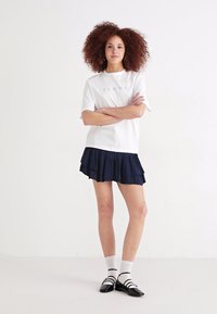 Biela tričko s krátkym rukávom s potlačou "FEMME", v kombinácii s navy sukňou s vrstvenými volánmi. Čierne topánky so bielymi ponožkami. Jednoduché pozadie.