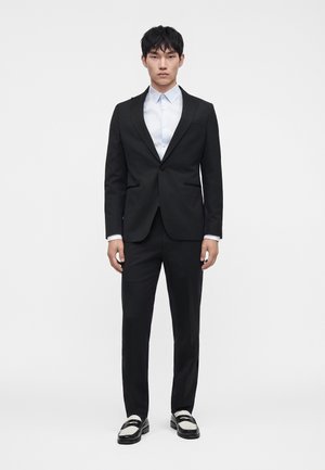 Homme portant un costume noir avec une chemise bleu clair et des mocassins en cuir noir et blanc, debout devant un fond blanc uni.