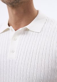 Polo de punto color blanco roto con un patrón de trenzado texturizado, tapeta de dos botones y un acabado de tela suave y lisa.