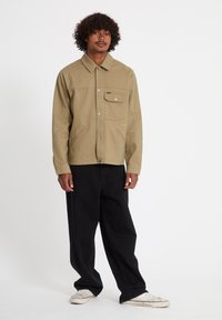 Homme aux cheveux bouclés portant une veste boutonnée beige, un pantalon noir ample et des baskets blanches, debout devant un fond blanc uni.