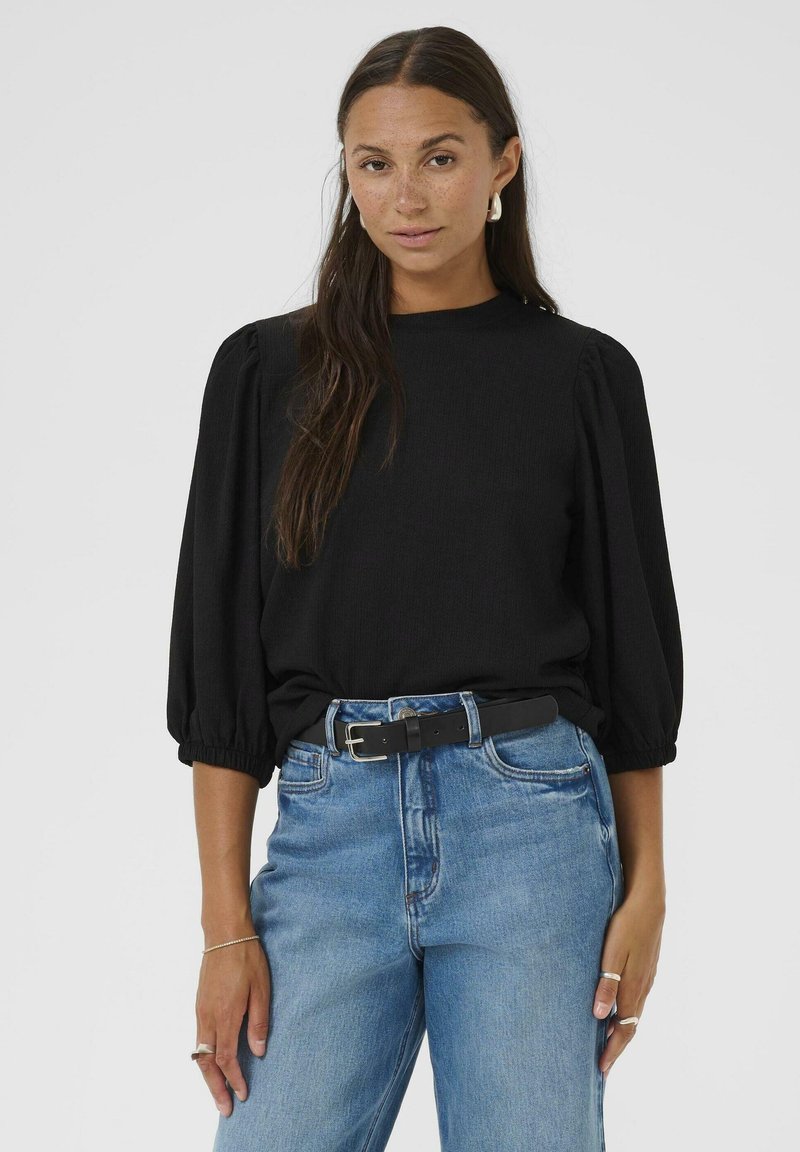 Haut noir à manches bouffantes en tissu texturé, associé à un jean taille haute bleu clair et une ceinture noire avec une boucle en argent.