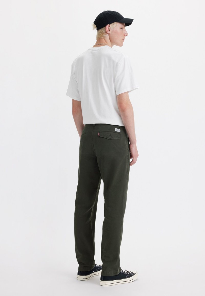 Camisa blanca de manga corta, pantalones verdes y gorra negra. Los pantalones tienen un bolsillo trasero con una etiqueta. Tela texturizada con corte recto.