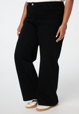 Jean noir à taille haute, à jambe large, avec cinq poches et une fermeture à bouton en métal. Tissu lisse avec une texture uniforme. Associé à des chaussures claires.