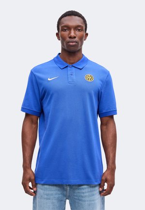 Uomo che indossa una polo Nike blu con un logo circolare giallo sul petto sinistro, abbinata a jeans azzurri, fermo in piedi.