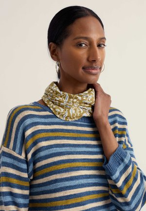Mujer de cabello oscuro que lleva pendientes de aro dorados, una bufanda floral verde alrededor del cuello y un suéter a rayas en azul, mostaza y crema, tocándose suavemente el cuello.