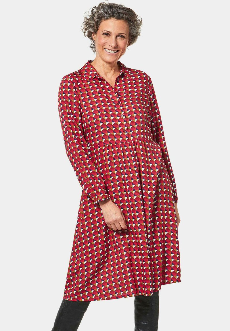 Vestito rosso con bottoni, caratterizzato da un motivo geometrico di triangoli in crema, giallo e blu. Maniche lunghe e vita arricciata.