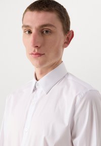 Weißes Hemd mit klassischem Kragen, glatter Textur und sauberem Finish. Verfügt über eine Frontleiste mit kleinen, runden Knöpfen.