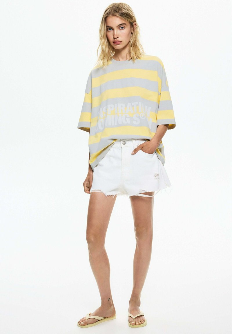 Grå og gulstribet oversized t-shirt med tekst, parret med hvide slidte denim shorts og beige flip-flops. Enkel design, afslappet pasform.