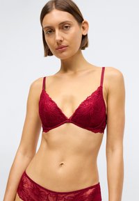 Γυναίκα με κοντά καστανά μαλλιά φορώντας σετ μαγιο σε μπορντό δαντέλα, απέναντι από έναν απλό αχνό φόντο.