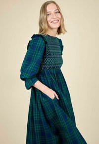 Femeie zâmbitoare cu păr blond, îmbrăcată într-o rochie în carouri verzi și albastre, cu un tiv brodat și detalii volă în umeri, cu mâna în buzunar.