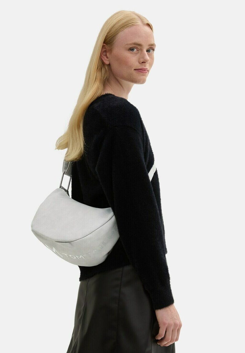 Weißer, weich strukturierter Beutel in runder Form mit geprägtem Logo. Modell trägt einen schwarzen, flauschigen Pullover und einen schwarzen, fließenden Rock, seitlicher Profilblick.