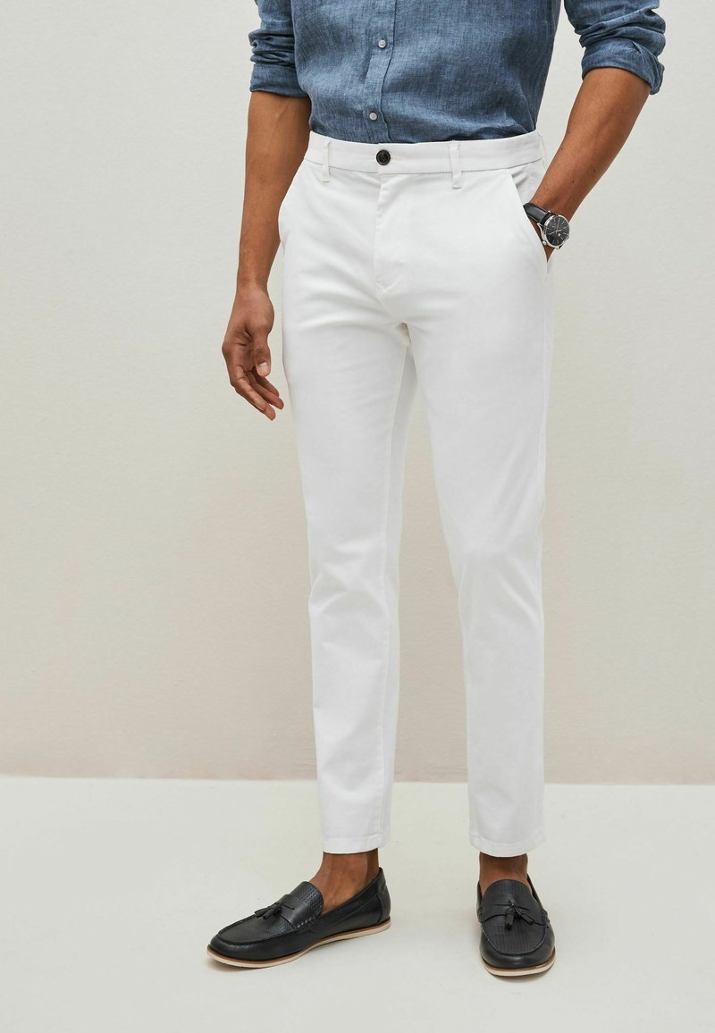 Pantaloni bianchi su misura con una vestibilità slim, realizzati in cotone. Presentano una chiusura con bottone e tasche. Abbinati a mocassini neri.