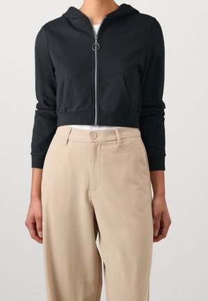 Kvinde iført en sort cropped hættetrøje med ringet træklynlås og beige højttaljede bukser, stående foran en ensfarvet baggrund.