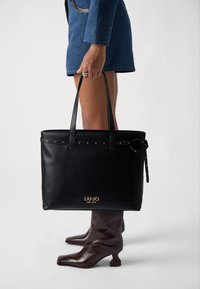 Bolso de tote de cuero negro con tachuelas doradas y una marca en relieve. Presenta asas dobles y una correa ajustable. Textura suave y diseño estructurado.