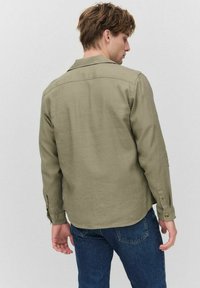 Olijfgroene langemouwenshirt met een puntkraag, achterpand en met knopen sluitende manchetten, gecombineerd met een blauwe spijkerbroek, gezien van achteren.