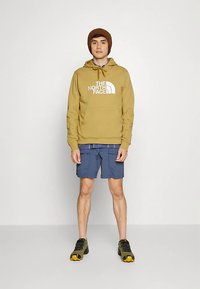 Jovem com gorro castanho, hoodie amarelo mostarda da The North Face, calções cargo azuis e sapatos de caminhada verdes, está de frente.