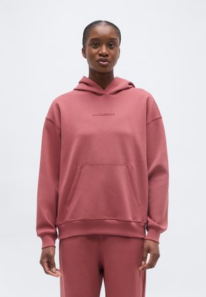 Naine, kes kannab summutatud roosa tooniga kapuutsiga pusa esitaskuga ja sobivaid pükse, seisab tühja heleda tausta ees.