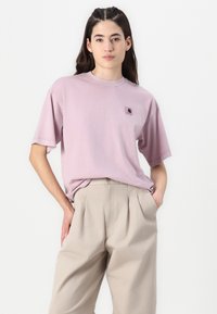T-shirt di cotone rosa chiaro con vestibilità oversize, maniche corte e un piccolo logo ricamato sul petto. Abbinata a pantaloni beige plissettati.