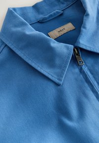 Gros plan sur une chemise à col bleu avec une fermeture éclair et une étiquette beige de marque portant l'inscription "NEXT" à l'intérieur de l'encolure.